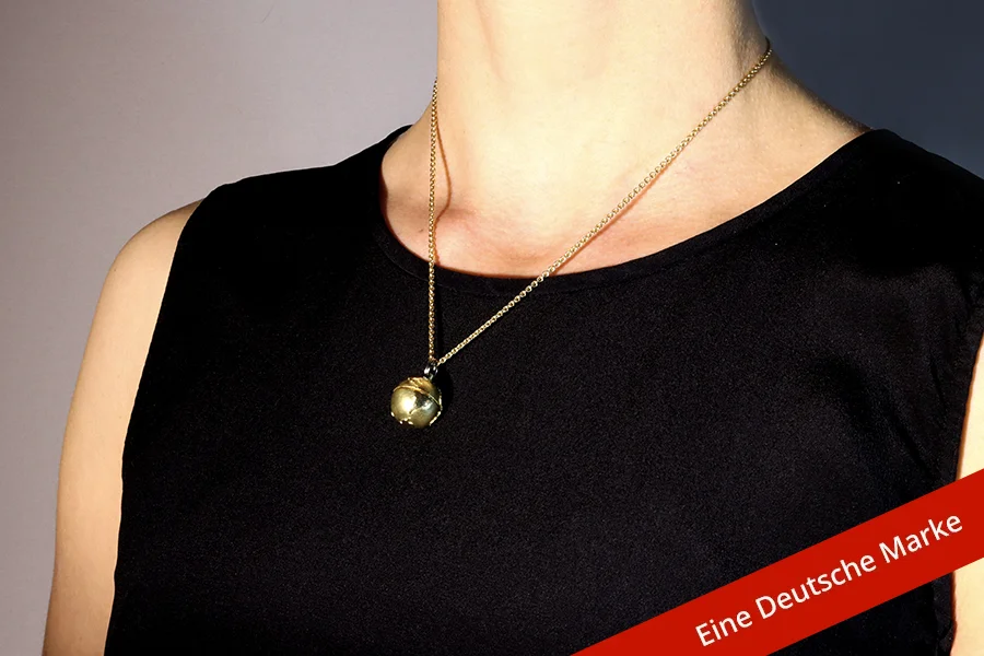 Eine Person in einem schwarzen Kleid trägt eine feine goldene Kette mit einem kugelförmigen, goldenen Medaillon oder Anhänger. Der Anhänger hat eine markante, gemusterte Textur. Ein rotes Banner in der unteren rechten Ecke trägt deutschen Text.