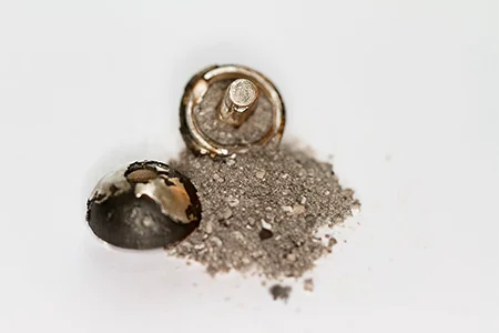 A close-up shows a small, open silver or metallic charm next to a small pile of fine grey ash, suggesting a memorial pendant designed to hold cremated remains.