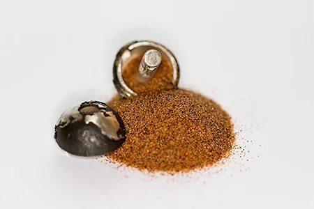 Eine Nahaufnahme zeigt einen kleinen, geöffneten silbernen oder metallischen Anhänger neben einem kleinen Haufen rötlich-braunen Sand. Eine Hälfte des Anhängers liegt auf der Seite, die andere ist leicht gekippt, mit einem sichtbaren Stift, der die Teile zusammenhält.