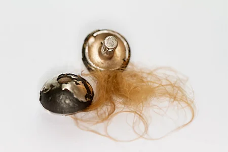 Eine Nahaufnahme zeigt zwei Hälften eines kleinen, geöffneten silbernen oder metallischen Anhängers. Daneben liegt ein kleiner Haarbüschel aus hellbraunem Haar, was darauf hindeutet, dass der Anhänger als Erinnerungsschmuck für das Haar eines geliebten Menschen gedacht ist.